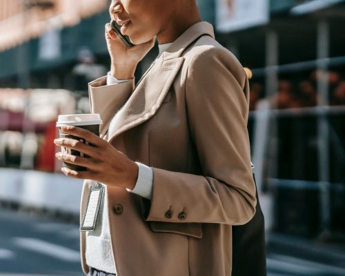 Side view of a young African American businesswoman talking on the phone with a takeaway coffee, walking outdoors.